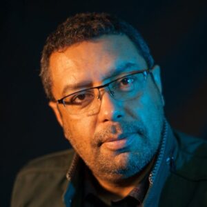 A thoughtful adult man wearing glasses, posing against a dark background.